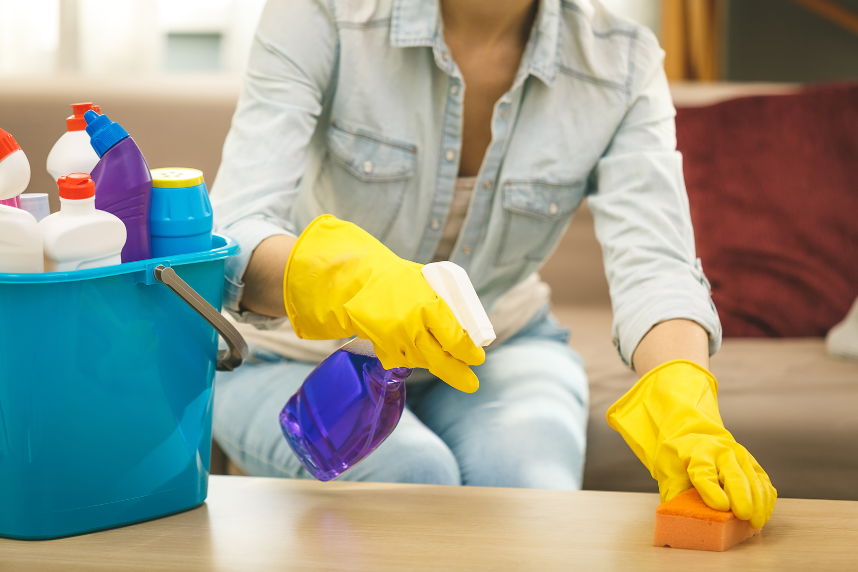 Woman in protective gloves is smiling and wiping dust using a spray and a duster while cleaning her house, close-up